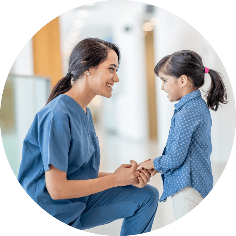 A medical professional in scrubs kneels to hold the hands of a young child.